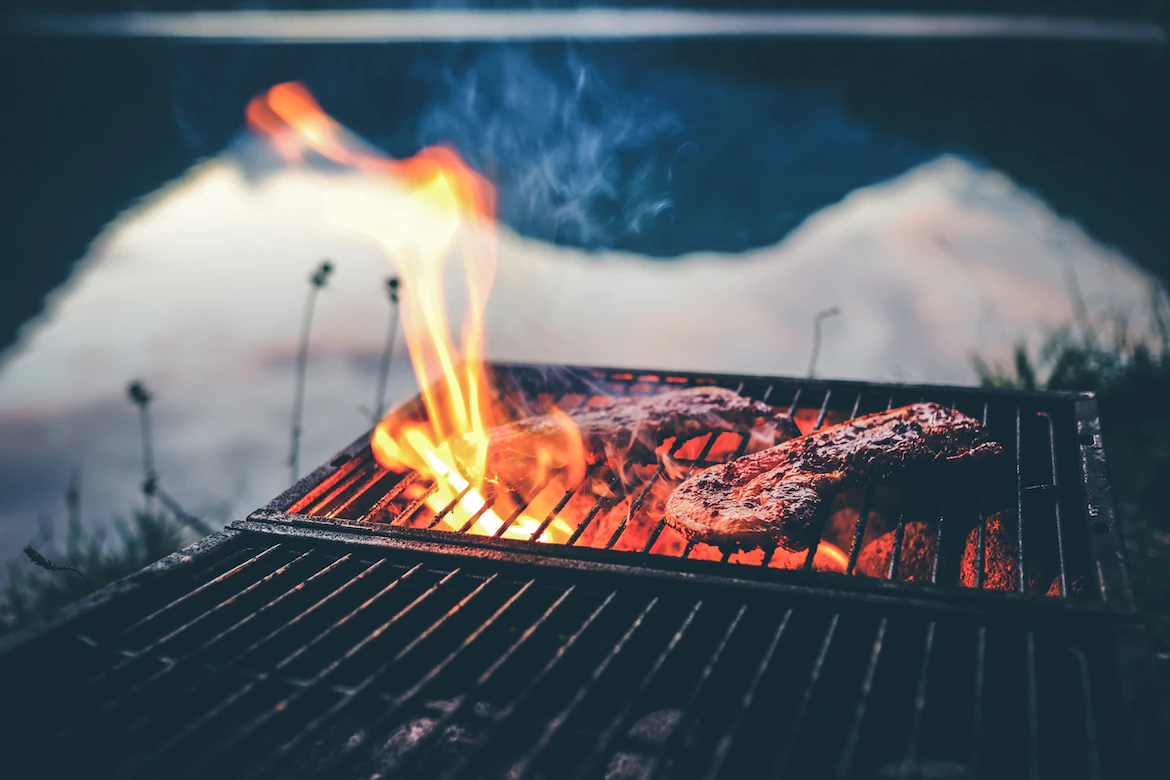 Steaks auf einem Grill mit offenen Flammen, im Freien neben einem Gewässer.